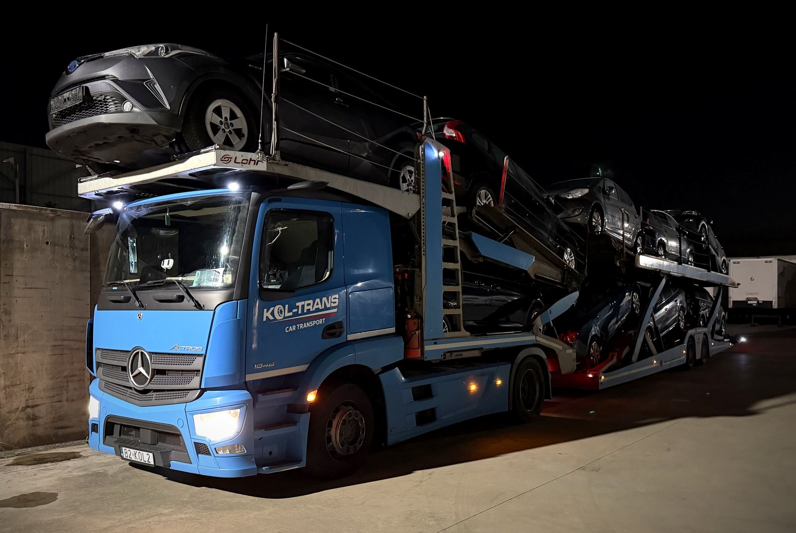 Niebieski autotransporter Mercedes-Benz Actros Kol-Trans z naczepą Lohr pełną samochodów, nocą na placu w Belgii