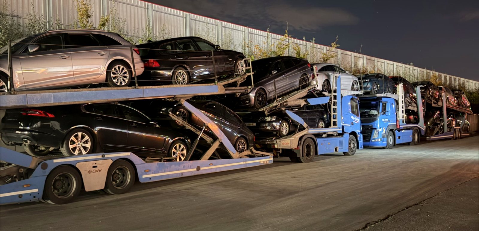 Dwa niebieskie autotransportery Kol-Trans – Mercedes-Benz Actros i Volvo FM z zabudową Lohr – załadowane samochodami, nocą na placu w Belgii